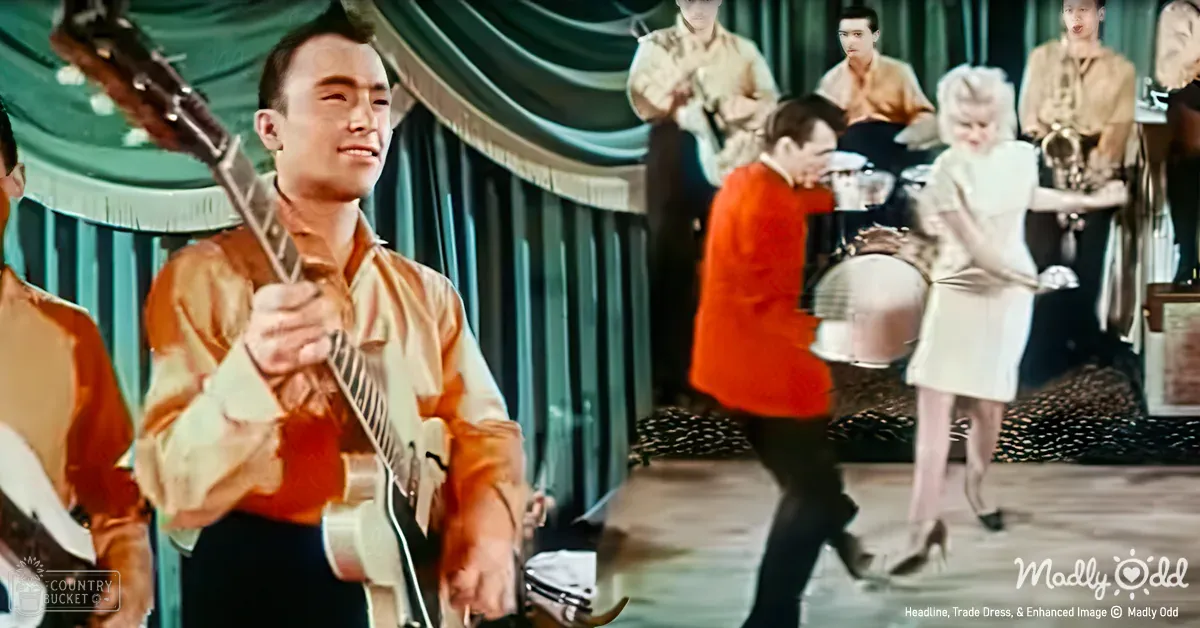 A young adult male musician plays guitar on stage during an evening performance, surrounded by other performers dressed in period clothing.