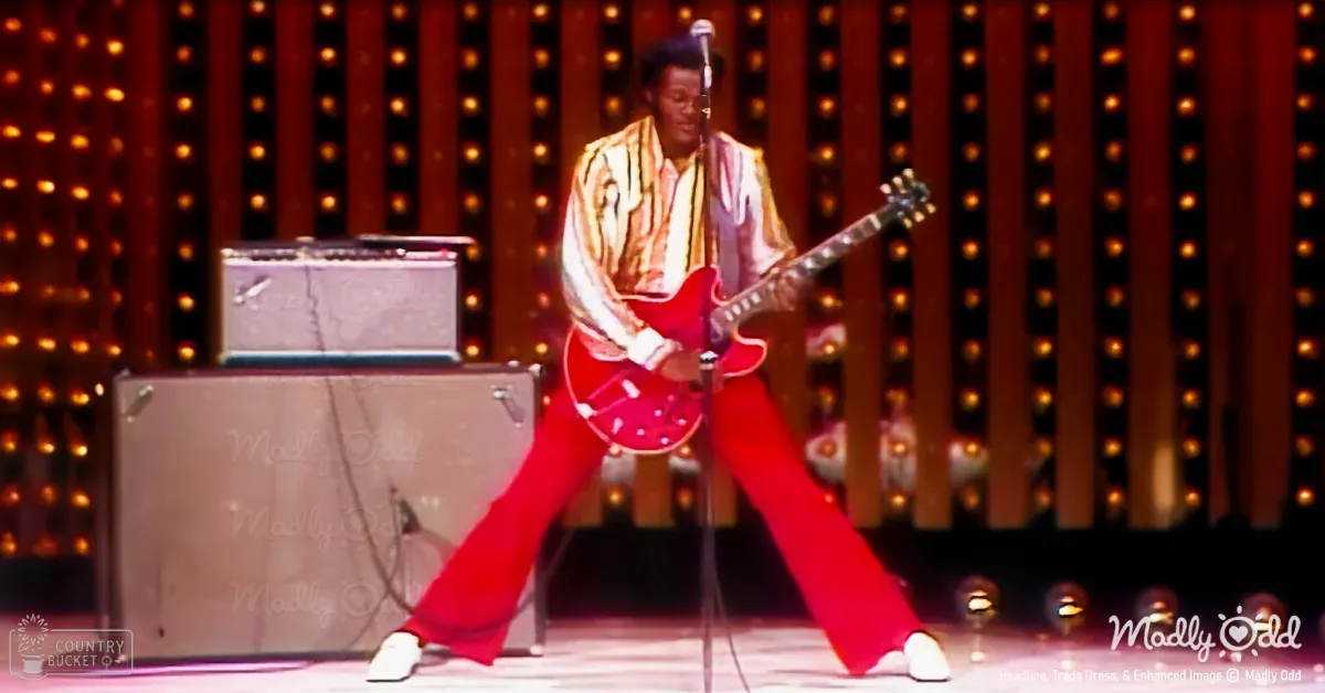 An adult male musician in a relaxed fit striped shirt and red pants performs at an indoor concert venue with two silver amplifiers, stage lights, and vertical background lines. The setting suggests it is nighttime or evening.