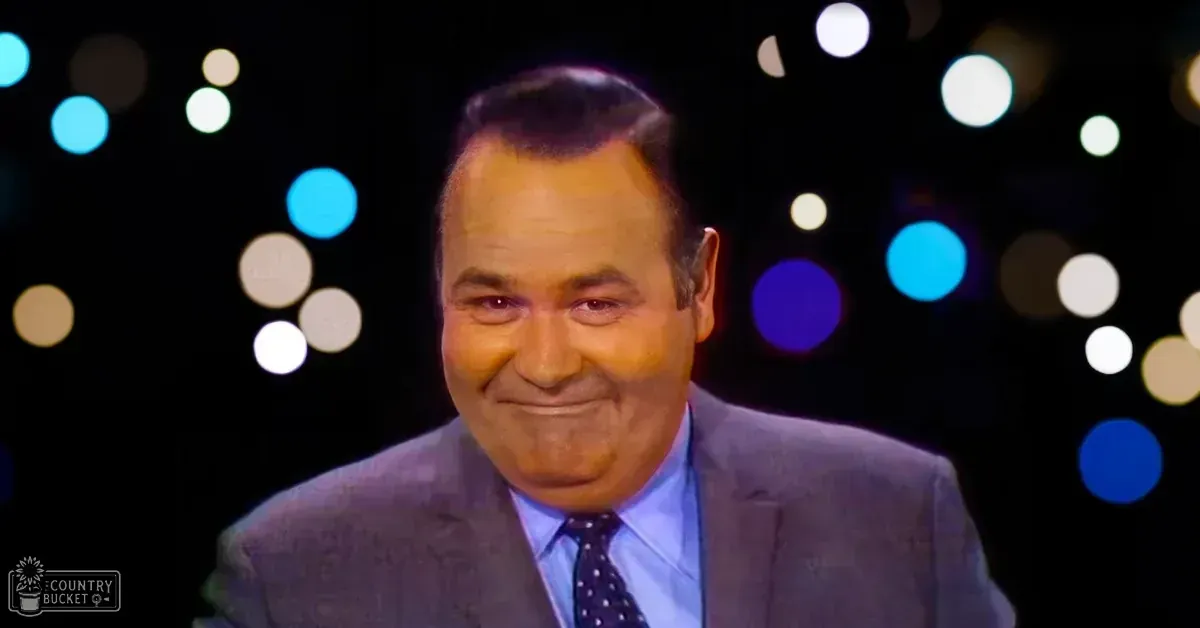An older adult male in a studio setting wearing a dark gray suit jacket with subtle patterns, light blue dress shirt, and a dark tie with small white dots. The scene suggests a formal or professional look from the mid-20th century.