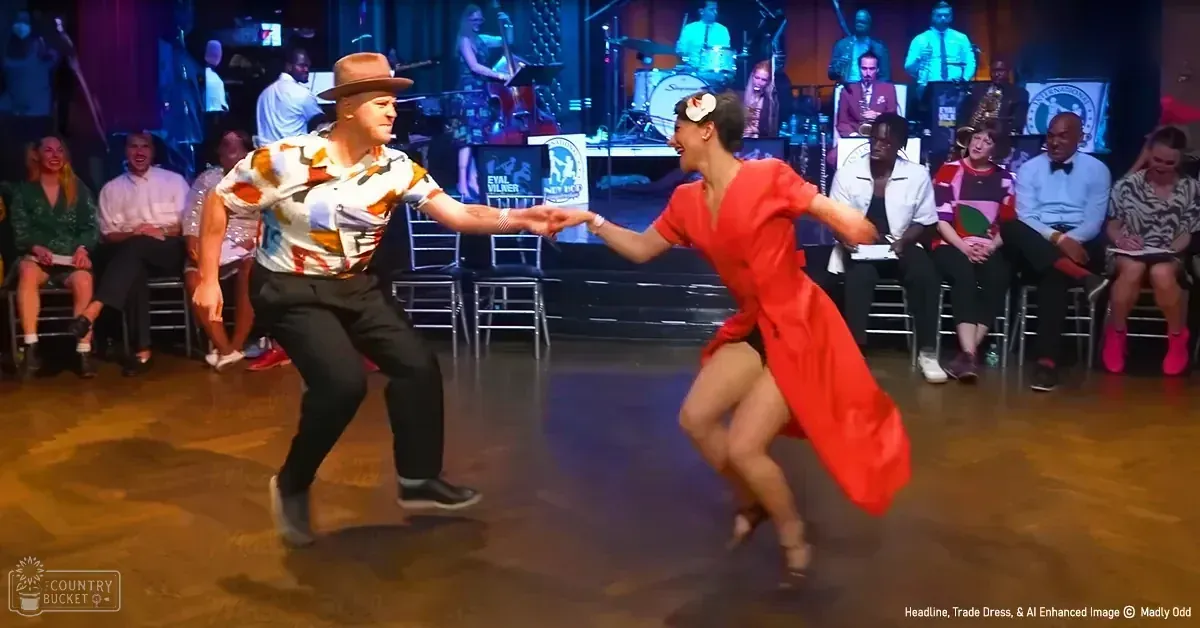 A dance duo performs on stage at an indoor event space with a seated audience. The male dancer wears a patterned shirt, dark trousers, and light sneakers; the female dancer is in a vibrant red dress with high heels.