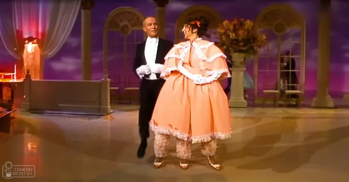 A couple dancing in an elegant indoor setting, possibly a grand ballroom. The male wears a black tuxedo and white gloves, while the female dons a long peach-colored dress with lace trim, also wearing white gloves.