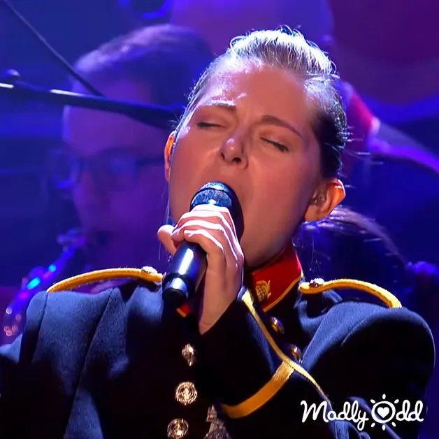 A young adult female sings into a microphone at what appears to be a performance setting with stage lighting and blurred figures in the background.