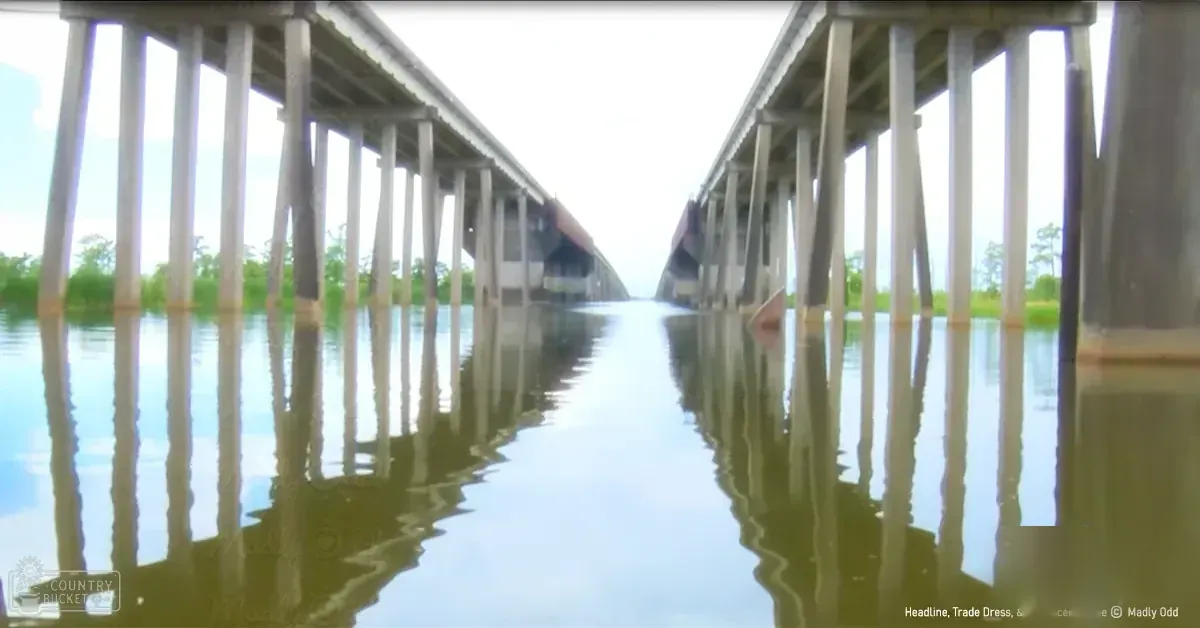 A long bridge spans a flooded area with greenery and partly cloudy sky, indicating daytime in spring or summer.