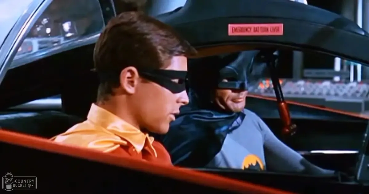 Young adult male in a classic utility suit with bat emblem, driving an iconic vehicle with futuristic interior details inside Gotham City on a bright daytime.