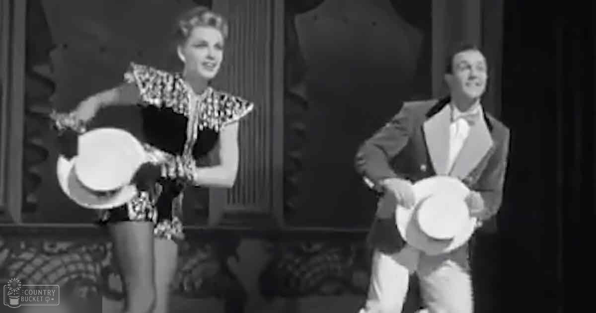 A theatrical performance scene from the early to mid-20th century with a young adult female holding a white hat and wearing a patterned dress, and a young adult male in a formal suit also holding a white hat, set against an indoor stage backdrop.