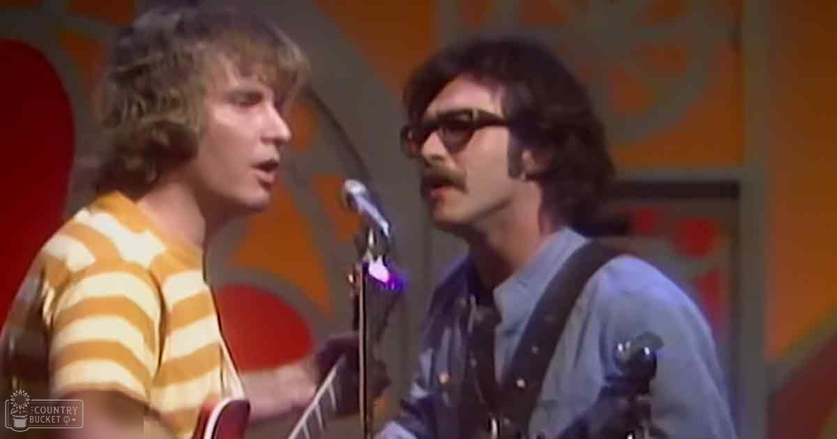A musical performance scene with two adult males, one in a yellow and white horizontally striped t-shirt and another in a blue long-sleeved shirt, performing or recording music on an indoor stage set with vibrant 1960s-70s era abstract designs.