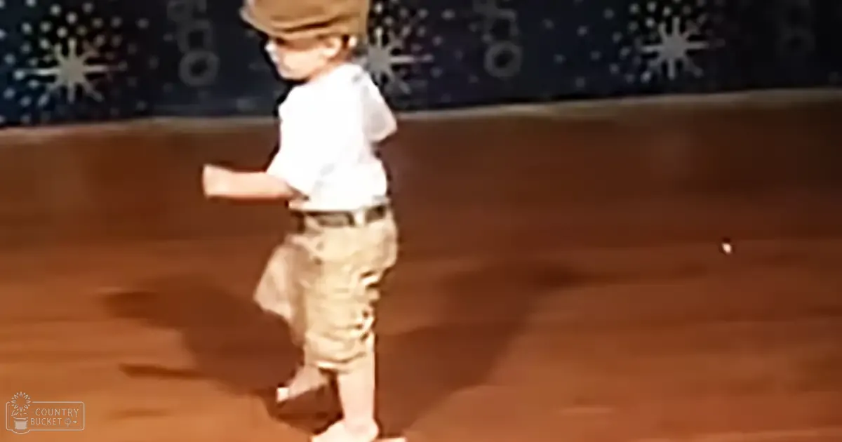 A toddler wearing a white short-sleeved shirt, khaki shorts or cargo pants, and a brown belt, standing indoors with star-like wall decorations.