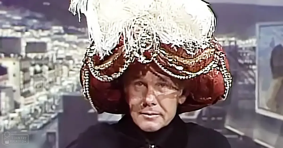 A middle-aged man wearing a large ornate red hat with white fringes and black and gold patterns stands indoors against an urban backdrop, suggesting cool weather.