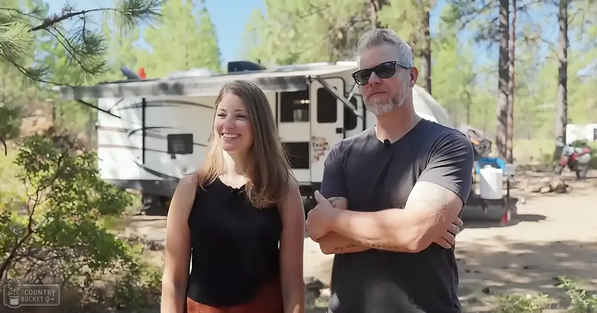 A middle-aged couple stands in front of an RV at a campground during summer with bright sunlight and green foliage.