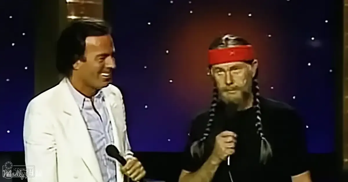 Indoor stage performance with two individuals, one wearing a white blazer over a patterned shirt and holding a microphone; another older adult male in a black t-shirt with a red headband, also holding microphones. Starry night sky backdrop suggests evening or nighttime setting.