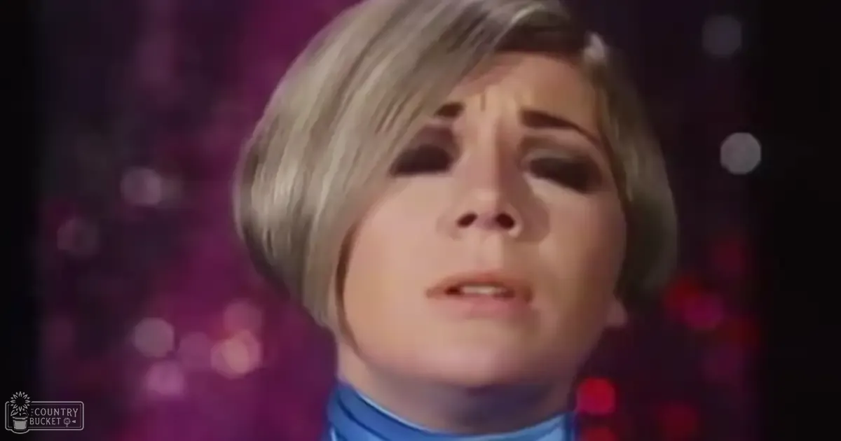 A middle-aged adult female wearing a high-necked, fitted turtleneck sweater in shades of blue indoors with blurred bokeh lights suggesting a stage or performance area.