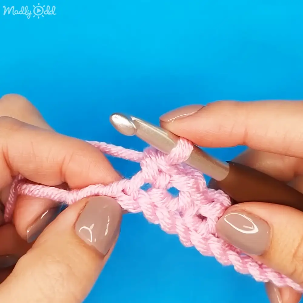 Close-up of hands crocheting with pink yarn against a blue backdrop, highlighting the craft and materials used in detail.