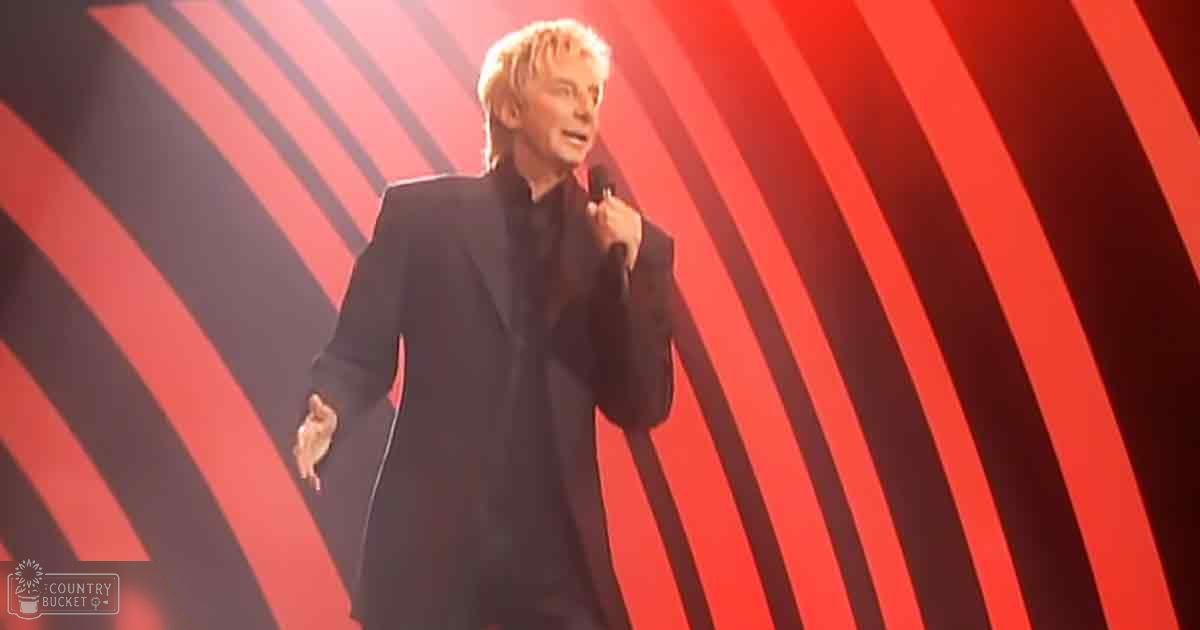 Adult male performer in a black suit jacket and trousers holding a microphone on stage with red and black curved lines in the background.