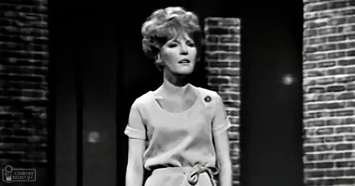 A young adult female in a casual, loose-fitting blouse stands on what appears to be a stage with brick walls and vertical bars, suggesting an urban or theatrical setting.