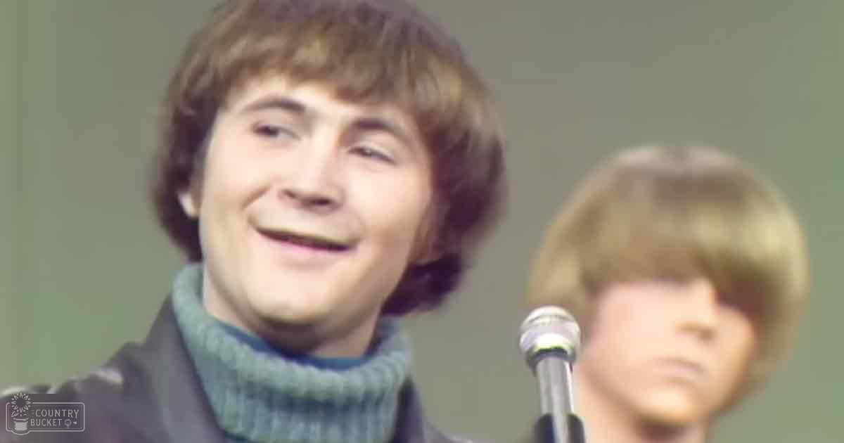 A young adult male in a dark jacket and teal turtleneck sweater stands in front of a microphone, likely at an indoor event. A child in a light-colored shirt is also present.