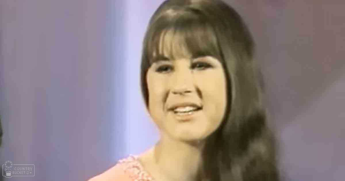 A young adult female wearing a light pink, lace-shouldered top with short sleeves indoors.