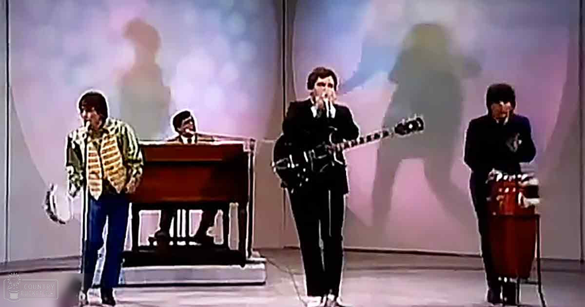 A musical performance in a studio setting with four adult male singers wearing dark suits, each positioned around a piano and microphones.