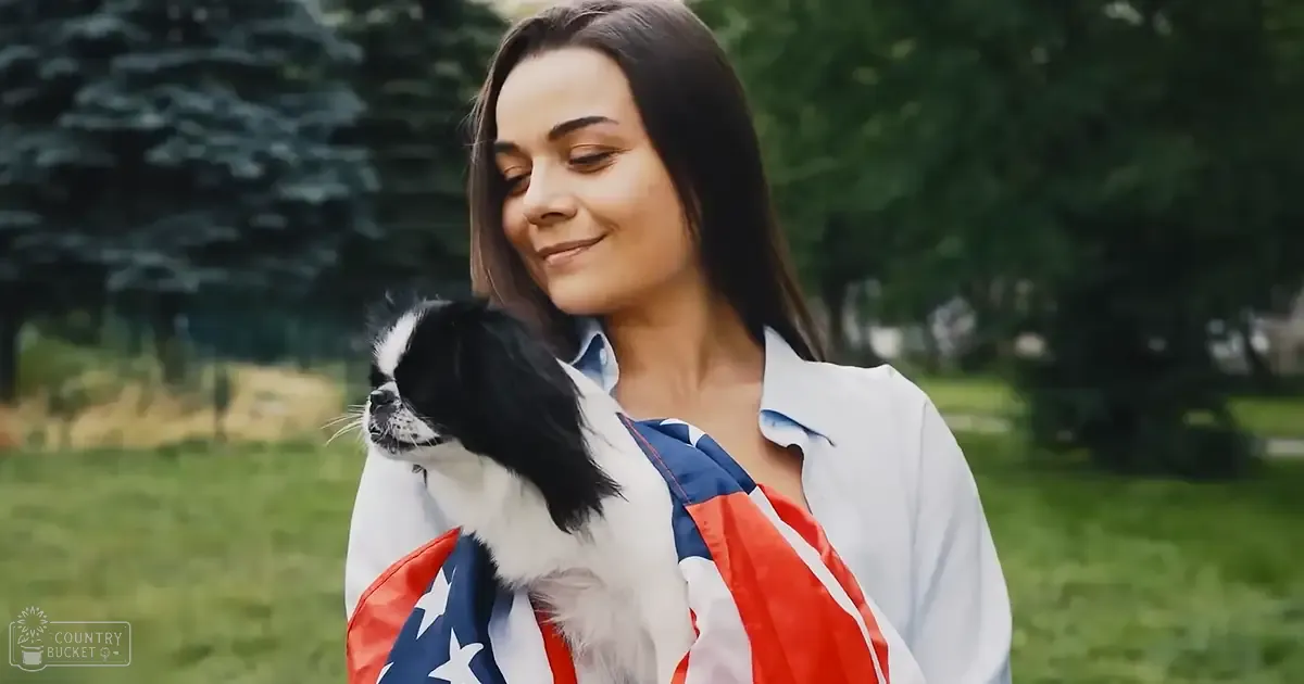 A young adult female wearing a long-sleeved button-up shirt with an American flag pattern in an outdoor park setting, accompanied by a small black and white dog resembling a Japanese Chin.