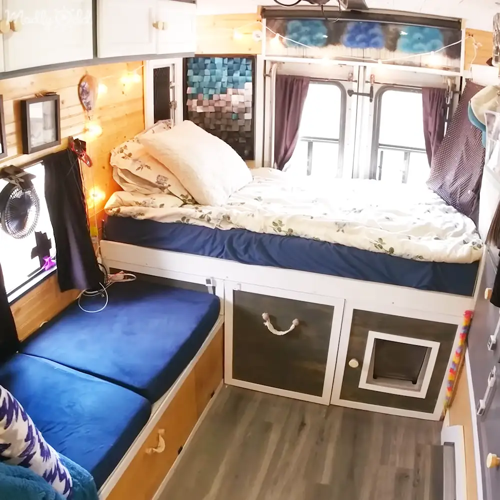 Compact bedroom setup in an RV or camper van with white floral bedding, blue mattress cover, string lights, and wooden paneling.