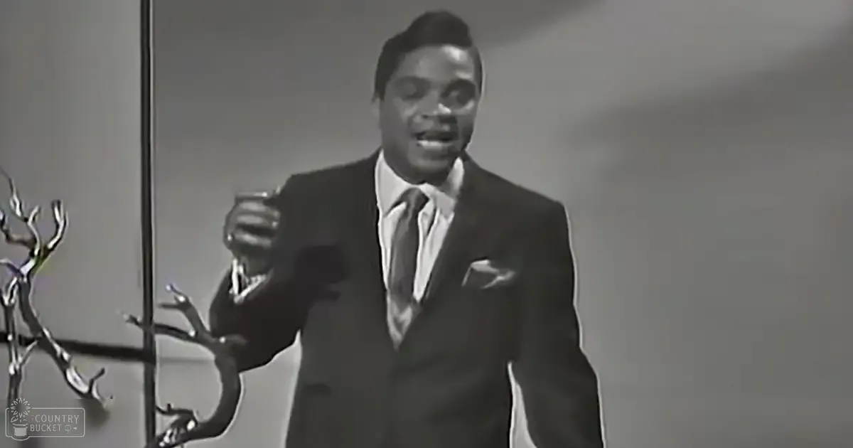 A man in an adult age band, wearing a dark suit and striped tie, stands on stage under focused lighting.