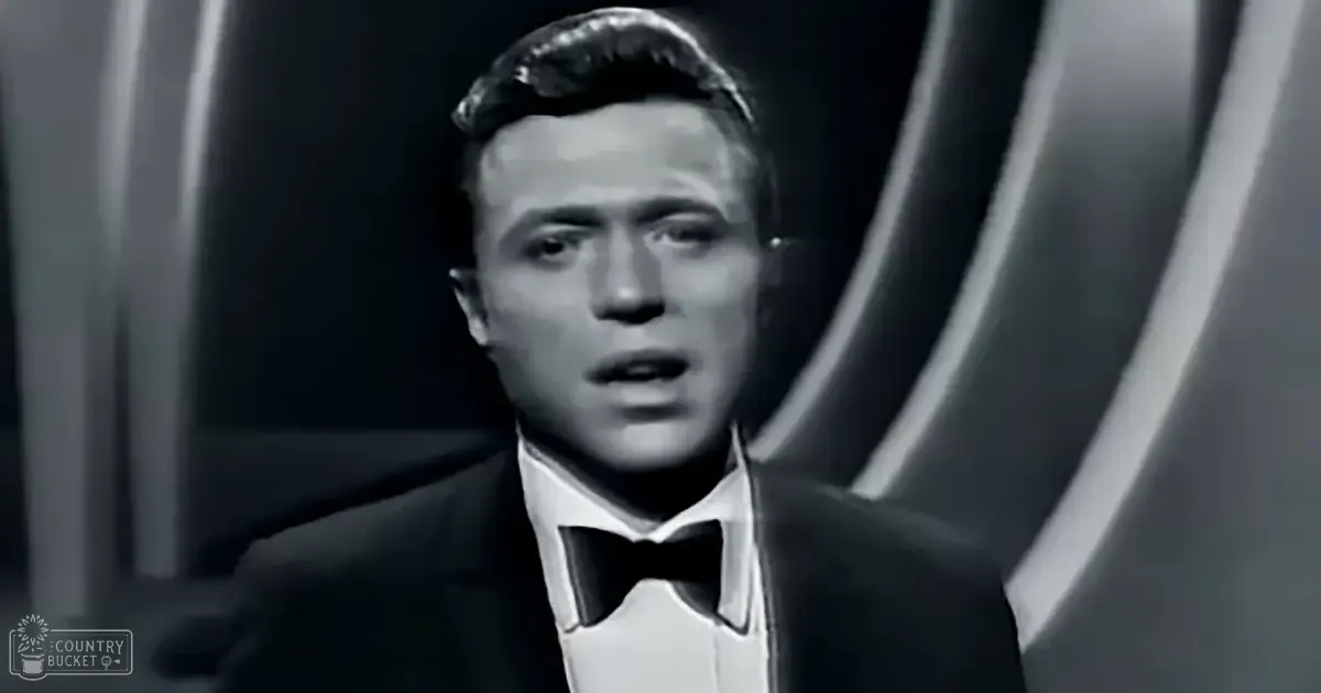 A young adult male wearing a classic tuxedo with a bow tie stands in an indoor setting illuminated by stage lights, with the text 'Madly Odd' visible.