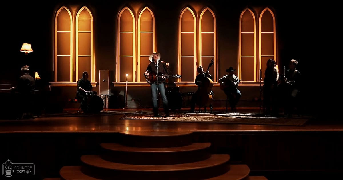 A musician in a church setting, performing with acoustic guitars and drums; adult male singer wearing a cowboy hat, dark shirt, light pants, and dark shoes.