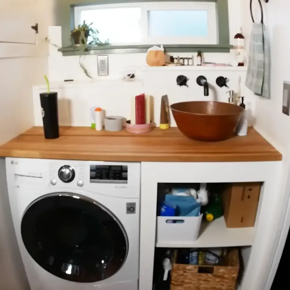 A home laundry area with a white LG washing machine, wooden countertop holding cleaning supplies and a black tumbler with green straw, under natural light from an unseen window.