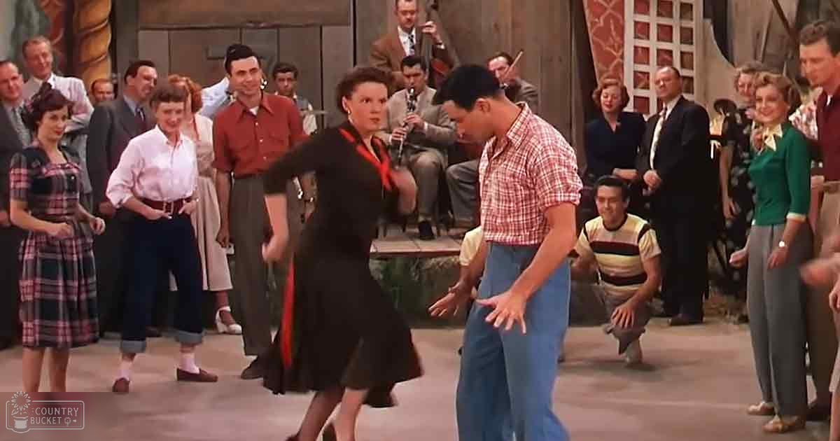A mid-20th-century dance scene set in an American town square with dancers in period attire performing in front of a stage with musical instruments, under natural daylight.