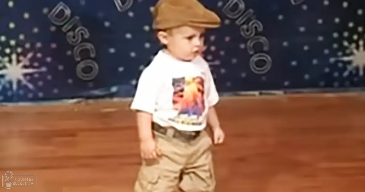 A toddler in an indoor setting, wearing a brown beret-style hat, white t-shirt with a character graphic, khaki pants; wall text 'DISCO' and starburst designs suggest a disco-themed party or event space.