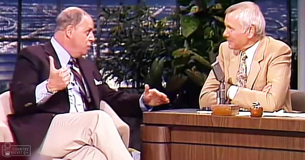 A talk show host and an older adult male guest in formal attire seated at a wooden desk with cityscape visible through large windows.
