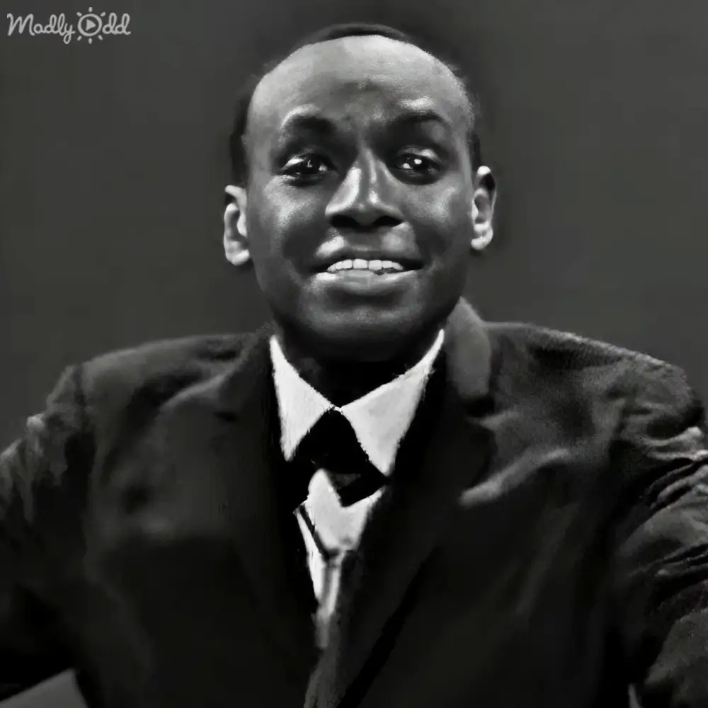 Adult male in a dark-colored suit jacket with a white shirt and tie against a plain indoor backdrop.