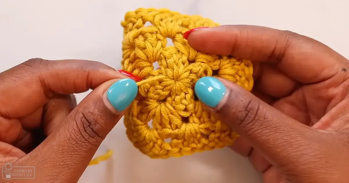 A crocheted flower with yellow thread, held by a person with blue and red nail polish in an indoor setting.