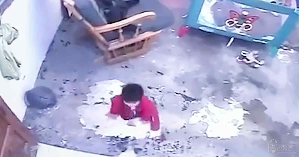 A toddler wearing a red long-sleeved shirt indoors with scattered items around, including a wooden rocking chair and toys on a small table.