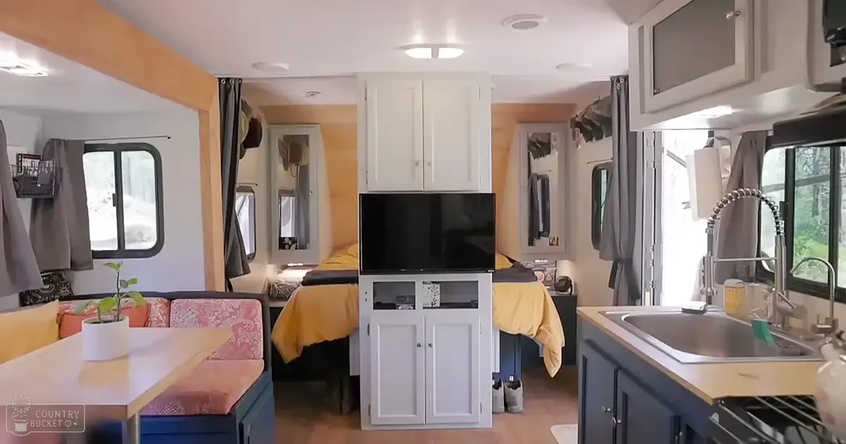 Compact RV interior with small kitchen, dining table, bed, TV, curtains, and potted plant in a residential setting during daytime.