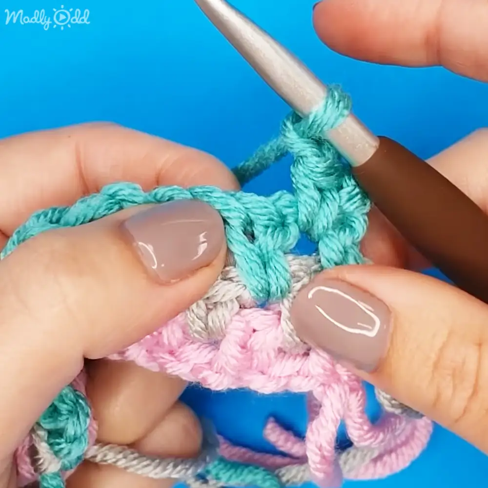A crochet project in progress with teal, pink, and gray yarns against a solid blue background indoors.