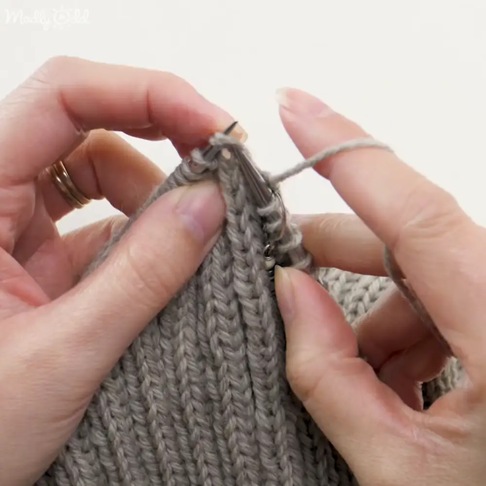 Close-up view of hands knitting with gray yarn against a plain white background, likely in an indoor setting