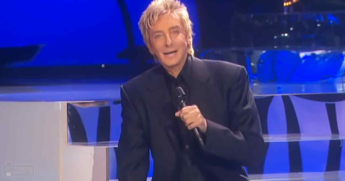 A middle-aged man in a dark suit jacket stands on what appears to be a stage or studio setup, holding a microphone with blue lighting and seating visible in the background.