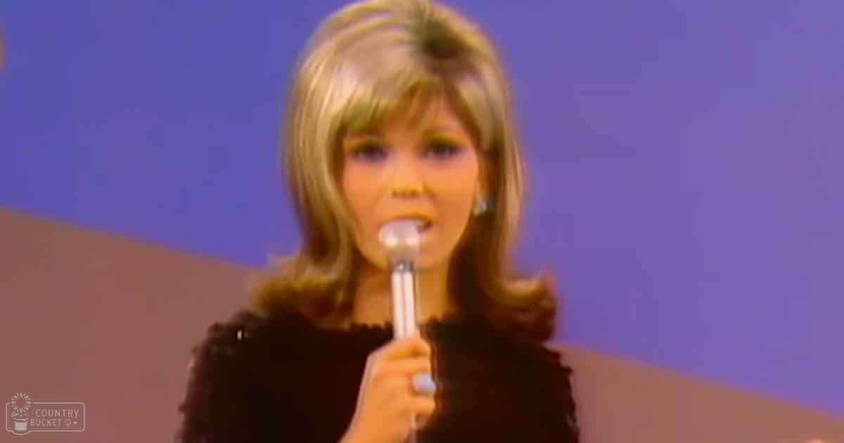 An adult female performer in an indoor setting, wearing a black textured garment with long sleeves, holding a microphone and labeled 'Madly Odd.'