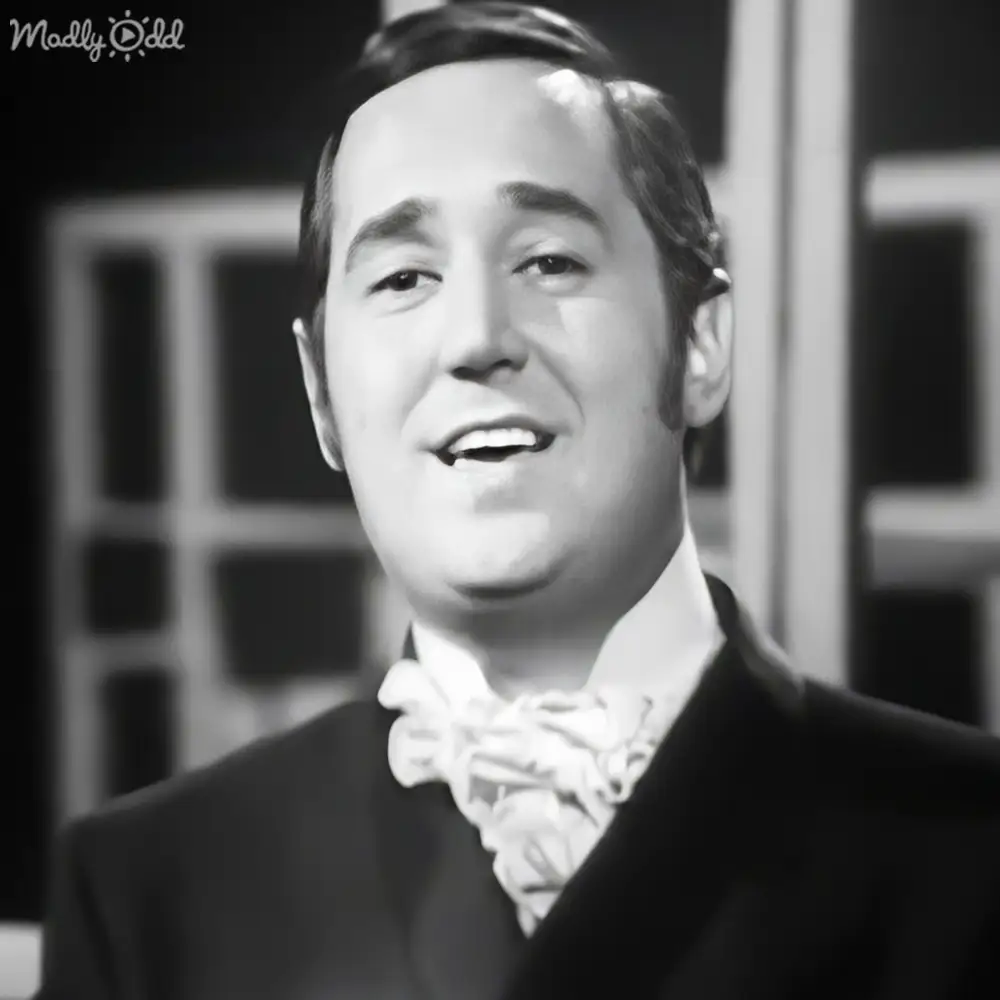 A middle-aged adult male wearing a formal outfit with a white cravat and dark jacket, set against an indoor background with soft lighting.
