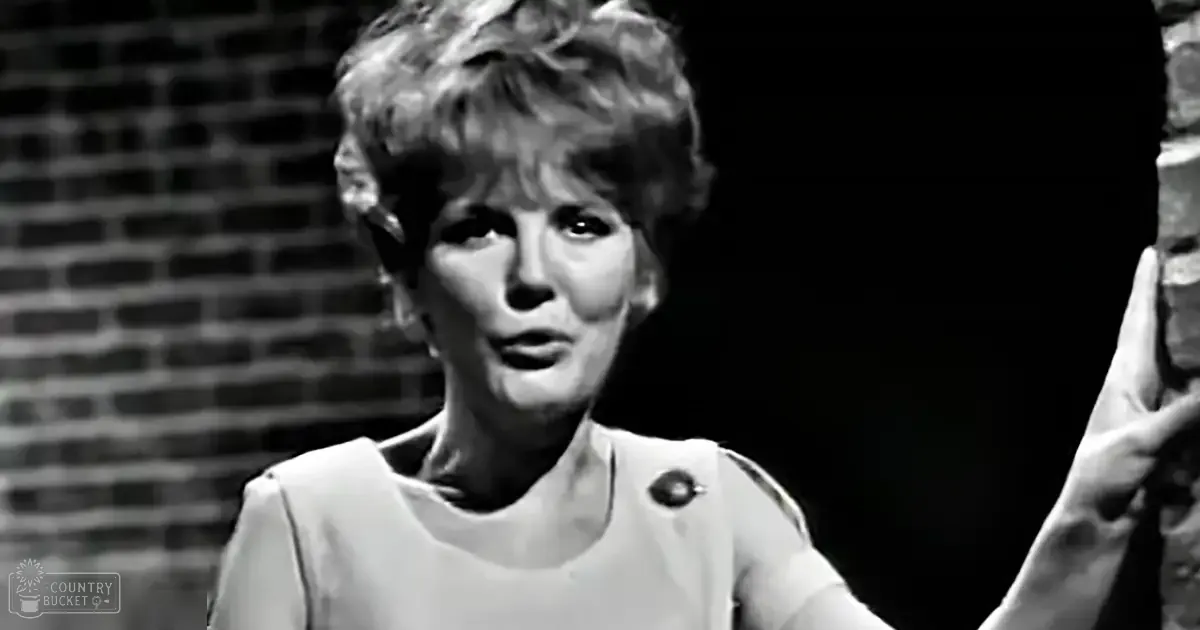 A middle-aged woman in a sleeveless top with a round neckline stands indoors against a textured brick wall backdrop under directional lighting, possibly during a performance or recording session.