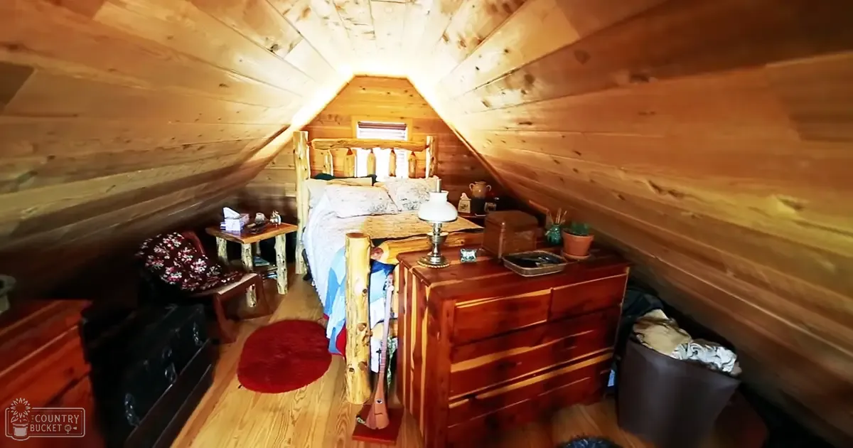 A cozy attic bedroom scene with sloped wooden walls, rustic furniture including a patterned mattress bed frame, floral-patterned chair, small round red rug on the floor, and natural daylight from a window at the top of the bed area.