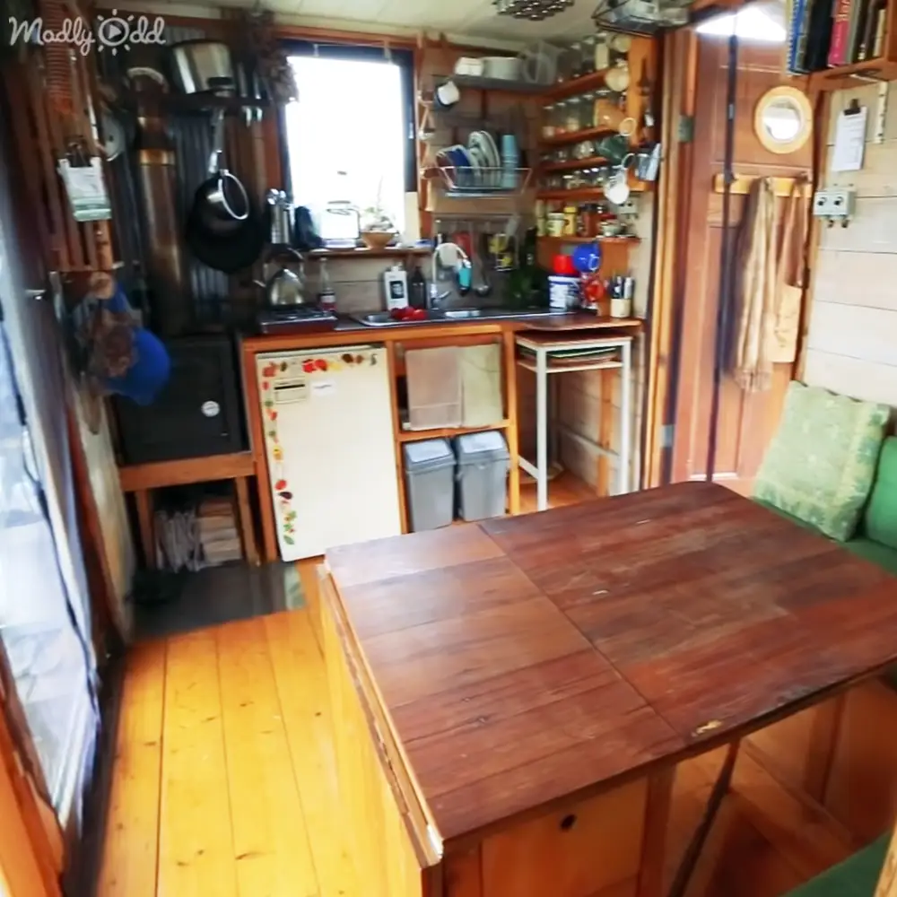 A cozy kitchen area with rustic wooden table, white refrigerator adorned with magnets, and natural light streaming through a window within a houseboat or small cabin.