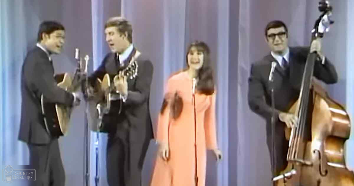 A musical performance with four adult individuals on stage, dressed in formal attire, holding acoustic guitar and double bass, set against a light blue curtain backdrop under natural or indoor lighting.