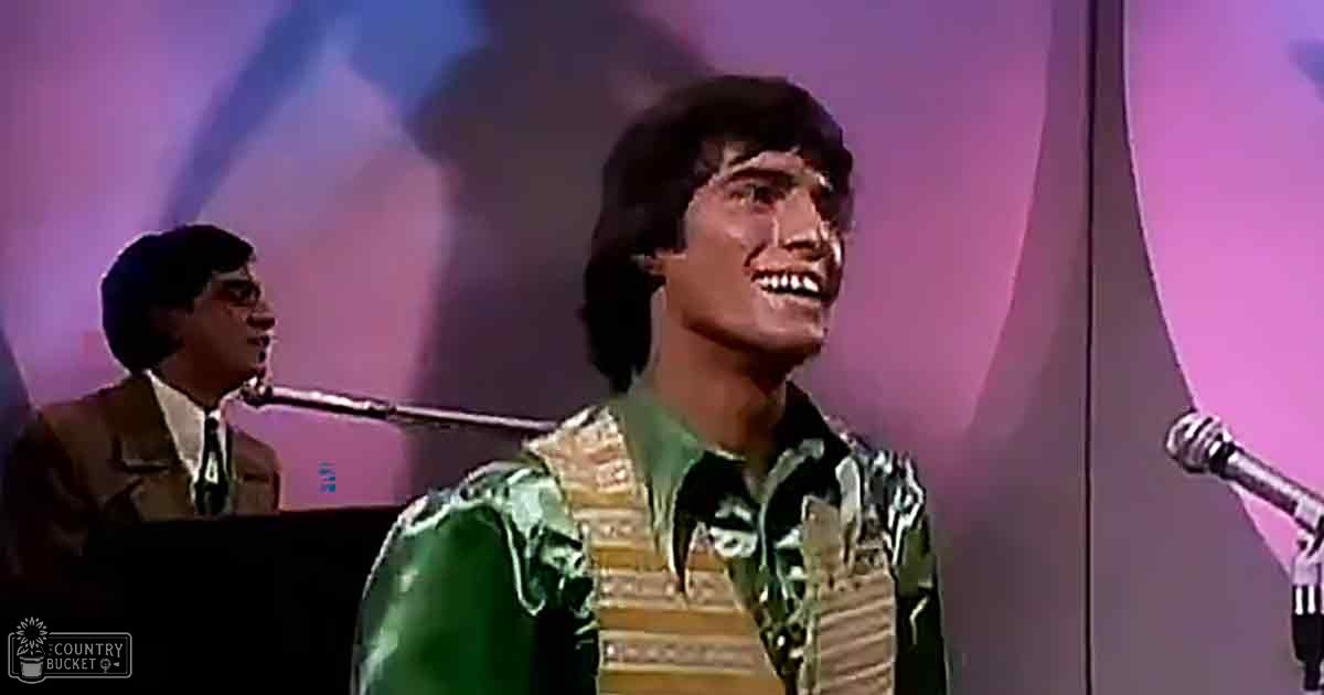 Young adult male singer in a green and yellow patterned shirt performing on stage with another young adult male in a brown suit, microphone stands visible.