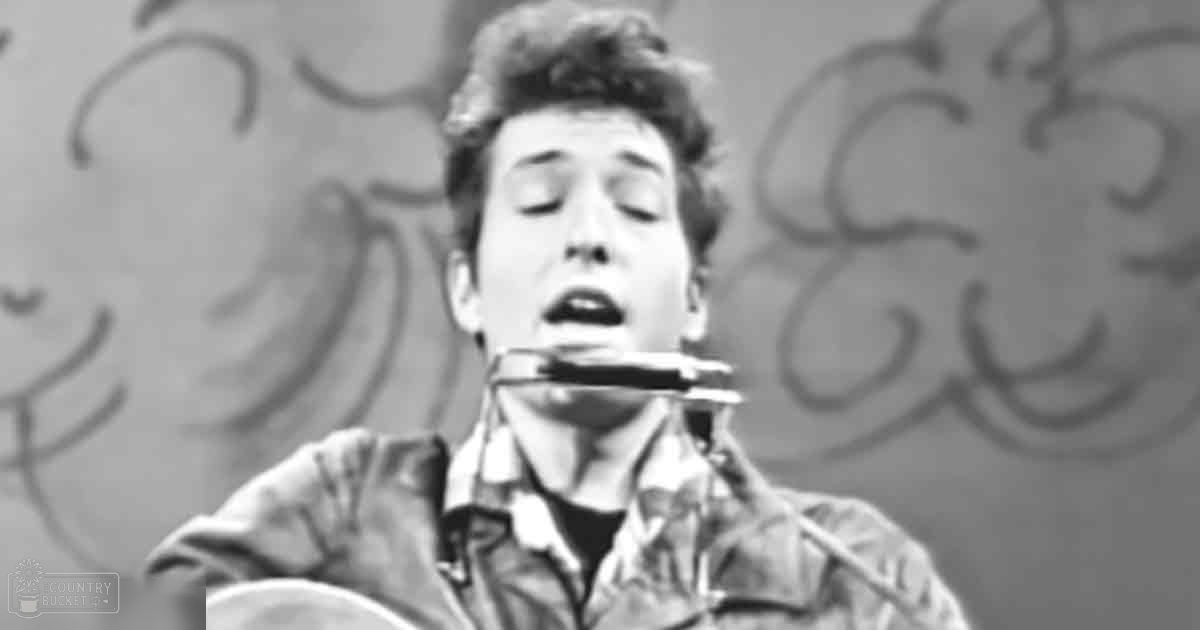A young adult male playing an acoustic guitar with a harmonica in his mouth, set against urban graffiti.