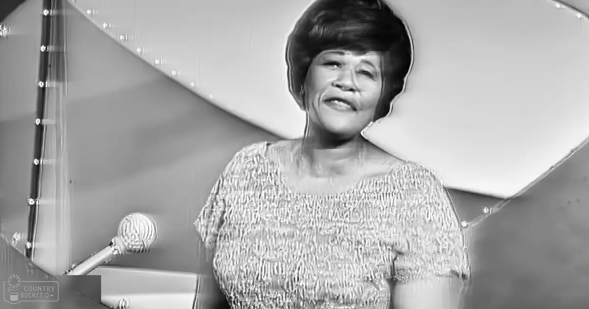 A middle-aged woman in a textured top with a round neckline stands indoors, surrounded by studio elements like a microphone and furniture.