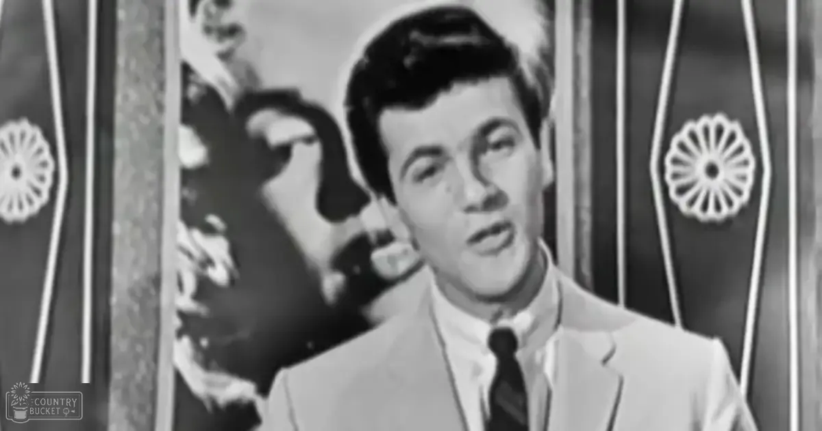 A young adult male wearing a light-colored suit jacket, white dress shirt, and dark tie stands against decorative floral panels in an indoor setting with even, bright lighting.