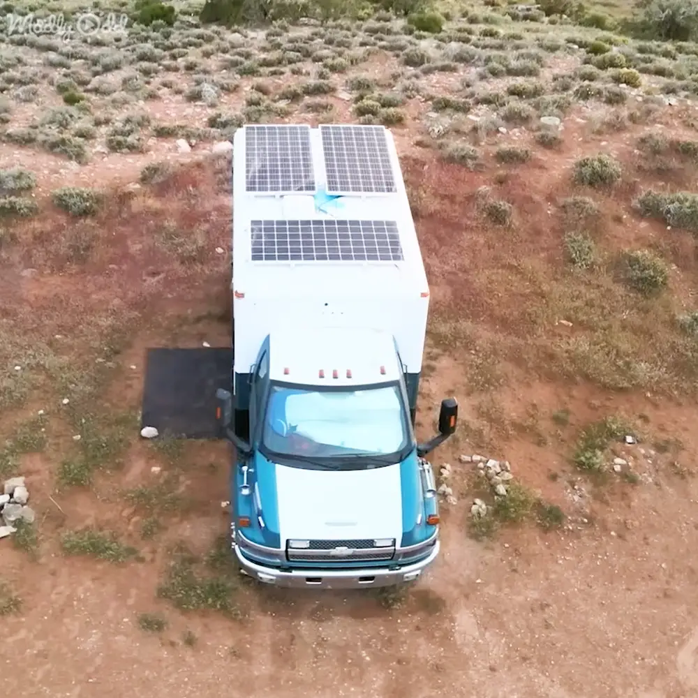 A white and blue van with solar panels parked in an open, arid landscape with dry reddish-brown earth and small shrubs.