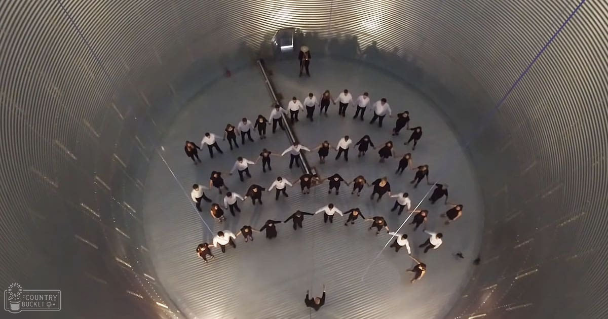 Young adults form a circle in an indoor setting with metallic, cylindrical walls; white long-sleeved shirts dominate the scene.