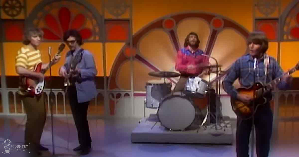 A musical performance indoors with young adult males in casual attire, surrounded by electric guitars, a drum set, and bright studio lighting typical of 1960s-1970s entertainment settings.